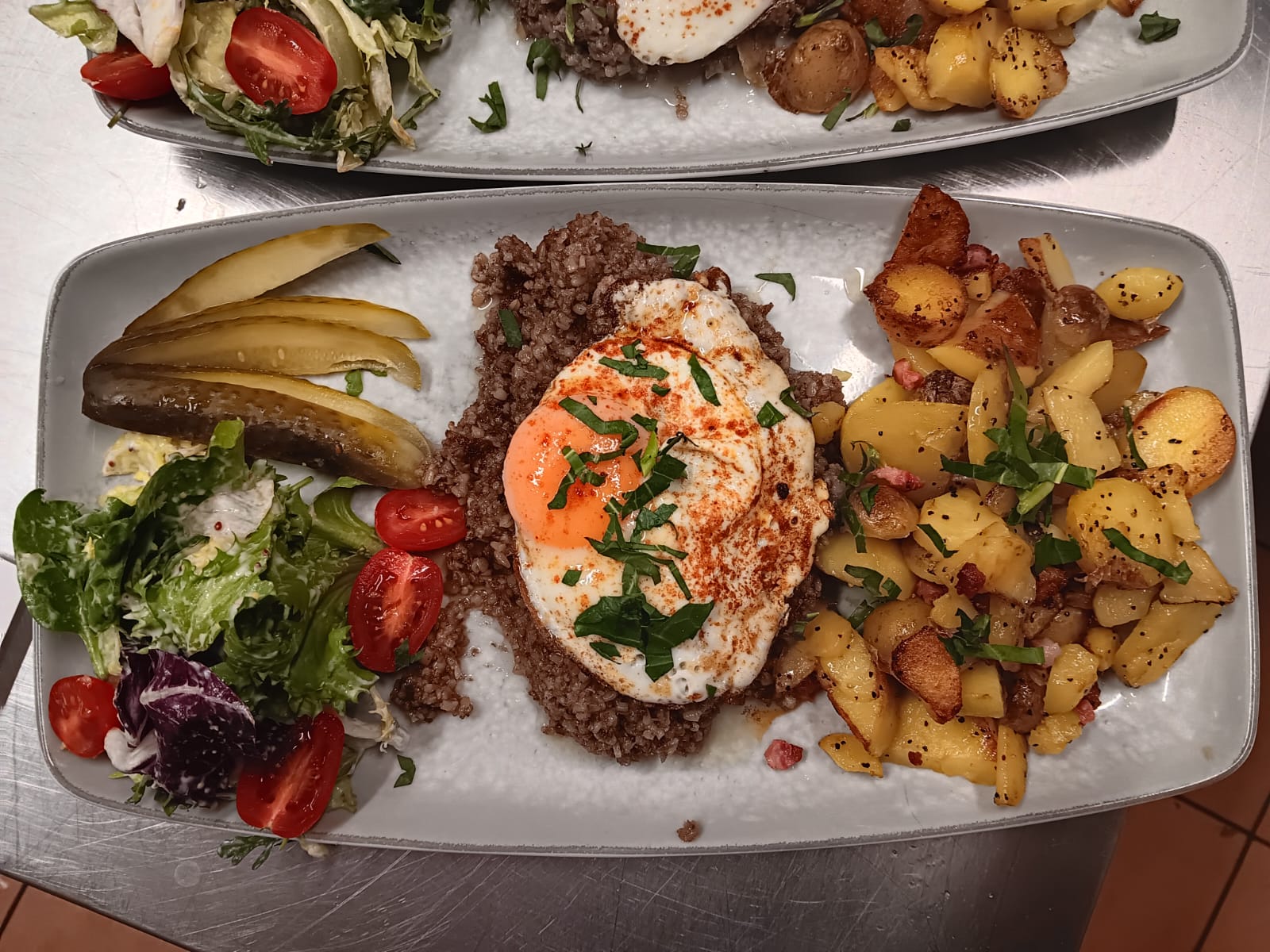 Ein Teller mit Spiegelei auf Hackfleisch, buntem Salat, Gewürzgurken und Bratkartoffeln